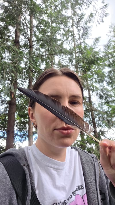 A woman is holding a dark feather in front of her face, obscuring her left eye and part of her nose. She is looking at the camera with her right eye visible. She is wearing a white t-shirt with text that appears to read "WELCOME TO WE RE" and a pink graphic, as well as a grey zip-up hoodie. She has a backpack strap across her chest. The background consists of tall trees with green foliage, suggesting an outdoor, possibly forested, environment. The lighting is overcast. The specific location in Portland, United States, cannot be determined from the provided image.