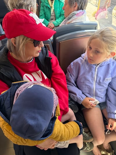 The photo captures a scene indoors, likely on public transport in Ostend, Belgium. A young girl in a light purple jacket sits near a woman with bare feet. Another child, wearing a blue hat and yellow jacket, rests on a woman's lap who is wearing a red shirt and baseball cap. An older woman in green sits nearby.