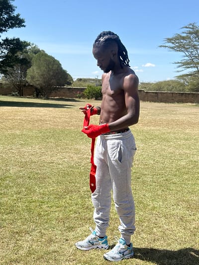 An adult male stands outdoors in a grassy area within Olkaria ward, Kenya, under a clear blue sky. He possesses dark skin, dreadlocks, and a beard, and is bare-chested, displaying a muscular build. He wears light grey ribbed sweatpants and white and light blue athletic shoes. The individual is actively wrapping his right hand with a long red hand wrap; his left hand is already wrapped in identical material. His attention is directed downwards towards his hands. The background includes a low, light-colored stone wall, multiple trees of varying sizes and foliage densities, and a expansive, short-cut grass field with areas of green and dry patches.