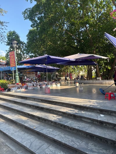 The image captures a scene in Lao Cai Ward, Vietnam. It features a row of stalls, each covered by a blue umbrella, lining a street next to a set of concrete stairs. The stalls are filled with various merchandise. A sign with red and yellow lettering, presumably indicating the street or area, is visible. Above the stalls, lush green trees with dense foliage provide a partial canopy, filtering the sunlight. The angle of the photograph suggests it was taken from the bottom of the stairs, looking towards the stalls and the street beyond.