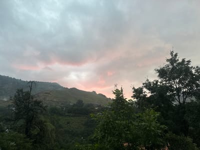The image depicts a cloudy sky with streaks of pink and orange, suggesting either sunrise or sunset. Below the sky, rolling hills are covered in lush green vegetation, interspersed with a few scattered houses and structures. The foreground is dominated by dark green trees, partially obscuring the view of the hills and houses. The overall atmosphere is serene and natural, with the soft hues in the sky adding a touch of beauty to the landscape. The time of day is likely early morning or late evening, given the sky's coloring. The weather appears overcast, but not stormy. The location is a rural or mountainous area, with the presence of houses indicating some form of human settlement within the natural environment. There is no text visible in the image.