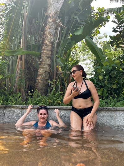 Two individuals are partially submerged in a body of water, positioned in what appears to be an outdoor setting with lush tropical foliage and a large tree trunk in the background. The person on the left, facing forward, is wearing a dark blue swimsuit and has their hands raised with fingers splayed. The person on the right, in a black bikini, is looking to their left and holding a mobile phone. They are both seated near the edge of the water, which is bordered by a stone wall. The environment suggests a warm climate.