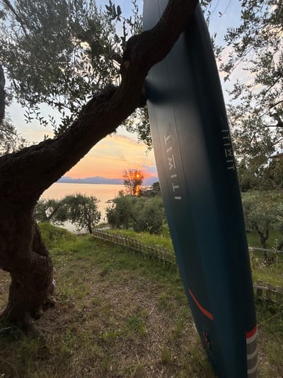 The image shows a paddleboard resting against a tree. The paddleboard is dark teal and has the "ITIWIT" logo printed on it in white letters. The scene is set outdoors, with a tree in the foreground and a body of water in the background. The sun is setting over the water, casting a warm glow on the sky and the landscape. The time of day appears to be sunset, and the weather is clear. The location is likely near a body of water, possibly a lake or the sea, given the presence of the paddleboard and the sunset over water. The image evokes a sense of peace and tranquility. There is also a wooden fence visible in the background.