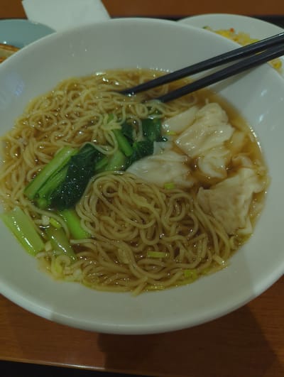A top-down, close-up shot captures a white ceramic bowl filled with wonton noodle soup, positioned on a wooden table. The soup contains thin, golden noodles, several translucent wontons, and bright green bok choy. Black chopsticks rest across the top of the bowl, poised to pick up food. The background is blurred, with hints of other dishes and what appears to be a napkin. The scene is set in an eatery in Narita, Japan, likely a restaurant offering traditional East Asian cuisine.