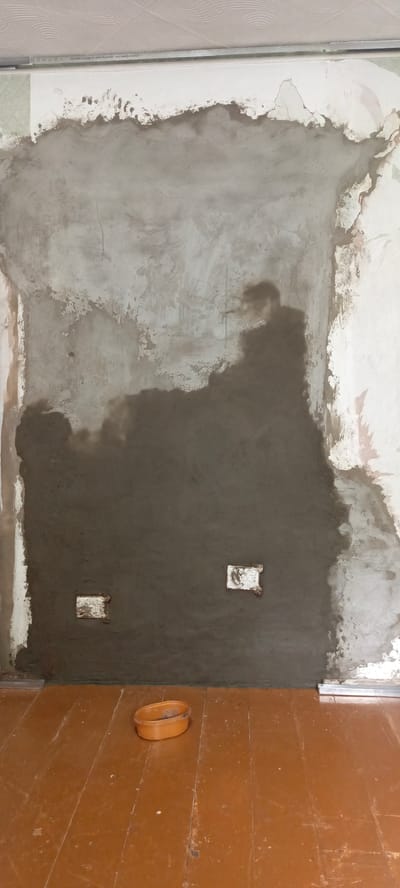 The image depicts an interior scene, likely a room undergoing renovation. A large section of the wall is covered in a dark gray plaster, partially obscuring older, lighter-colored plaster and wallpaper remnants. Two white electrical boxes are visible embedded in the plastered wall. The floor is made of worn, brown wooden planks. In the foreground, on the floor, sits an orange plastic container. A silver metal trim piece is visible along the bottom edge of the wall, near the floor. The ceiling, partially visible at the top, appears to be made of textured panels. The overall impression is of a work-in-progress, with materials and evidence of construction present.