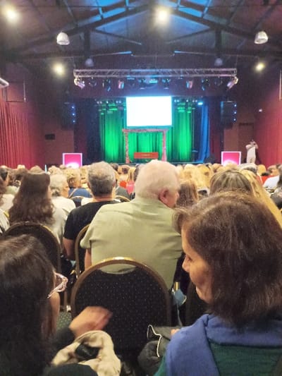 The image shows an audience seated in a theater venue in General Pacheco, Argentina (latitude: -34.457, longitude: -58.633). The audience is facing a stage with a large blank screen and a green curtain behind it. A single person is standing on the stage, facing away from the audience. There is a red curtain to the left side of the stage, and a similar red curtain to the right side of the stage. The room has a high ceiling with exposed beams, and there are multiple rows of seats for the audience. There are numerous people in the audience, and the majority are focused on the stage. Some people are seated, while others are standing or moving. Some of the audience members have their hands in their laps, others are talking with their neighbors, and one person is looking off to the side.