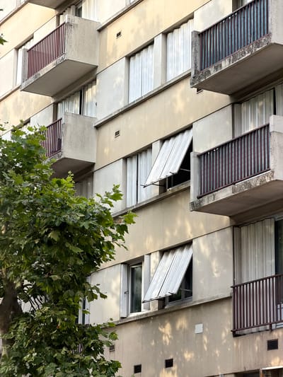 A low-angle, full shot captures the exterior of a beige, mid-rise apartment building in Maisons-Alfort, France, with sunlight casting dappled shadows from an unseen tree. The building features multiple balconies with dark red railings, some of which are partially obscured by the tree's green foliage. The windows have white, slatted shutters that are partially open on some levels, allowing glimpses of curtains inside. Small, dark rectangular vents are visible on the building's facade, and a white square electrical box is also present. The concrete elements of the balconies show signs of weathering and staining. The scene is devoid of any human presence.