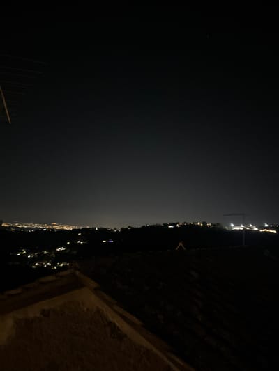 The image shows a night view of Castelnuovo di Porto, Italy, captured from an elevated position, possibly a building or hillside overlooking the town. The foreground is dark and out of focus, with a textured surface (possibly a wall or rooftop) visible in the bottom right. The left side of the frame displays the night sky, with minimal visible stars. The lights of Castelnuovo di Porto stretch horizontally across the middle of the image. A crane structure can be seen among the lights. A dark slope with some foliage outlines the town.