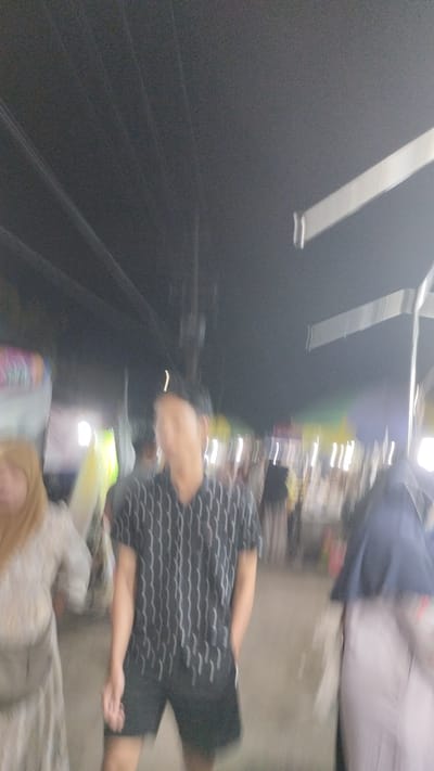 The image depicts a street scene at night, likely in Sambungrejo, Indonesia, characterized by the presence of people and various stalls or vendors, suggesting a market or festival atmosphere. The lighting is dim and appears to be artificial, with some sources casting bright, elongated streaks due to motion blur.
The central subject is a person wearing a short-sleeved shirt with a vertical striped pattern in black and white, and black shorts. To the left of this individual, another person is partially visible, wearing a light-colored outfit and a headscarf. To the right, another person is seen wearing a dark hijab and a striped skirt. The overall image suffers from significant motion blur, making it difficult to discern fine details or facial expressions.
In the background, there are indications of stalls or structures with lights, and the presence of other blurred figures suggest a gathering of people. Overhead, what appear to be electrical wires are visible against the dark sky. Two white, elongated shapes, possibly part of a structure or signage, are also prominent in the upper portion of the image. The time of day is clearly night, and there are no immediate cues about the weather, though the low lighting might suggest it is not raining. The scene conveys a sense of activity and public gathering, typical of a night market or local event. No visible text is discernible due to the blurriness.