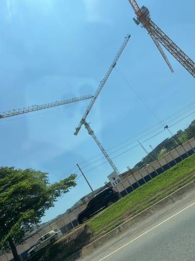 This image captures a construction scene in Jiru, Nigeria, under a clear, bright blue sky. Four yellow lattice-boom tower cranes are prominently featured against the sky, indicating active construction. One crane is in the upper right, displaying its machinery housing and operator's cab; another extends horizontally across the mid-left; a third extends downwards in the middle-right; and a fourth is partially visible on the far left. Below the cranes, a segmented construction fence with light-colored panels and a dark base separates a construction site from a paved road. Behind the fence, a white sign with a red circular logo and the black text "ITB" is partially visible. Further back, a rising landmass or hill shows visible concrete structures, suggesting ongoing building development. A strip of vibrant green grass runs along the roadside, bordered by a concrete curb. A paved road with a white line marking occupies the lower right quadrant. A dark-colored SUV is parked on the grassy verge, partially obscuring the ITB sign, and a lighter-colored sedan is visible further down the road to the left, partially hidden by a large, dense green-leafed tree. Utility poles and power lines traverse the upper and mid-ground. No individuals are discernible in the scene. The image is captured with a noticeable upward and counter-clockwise tilt.