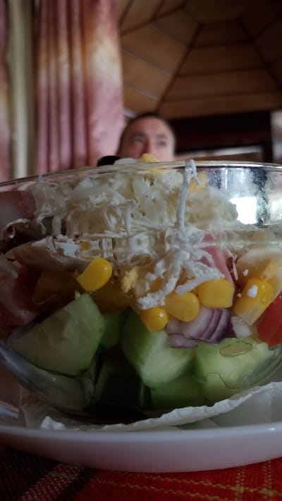 A close-up, eye-level shot captures a glass bowl filled with a colorful salad. The salad contains chopped cucumbers, corn kernels, red onion slices, and what appears to be shredded cheese and possibly boiled eggs. A person's face is visible in the blurred background, looking upwards. The setting appears to be indoors, possibly a restaurant, with a wooden ceiling structure visible above. A portion of a red and yellow checkered tablecloth is in the foreground, suggesting the bowl is placed on a table. The image is focused on the salad, with the background elements out of focus. The location in Pevtsite, Bulgaria is not explicitly visible in the image itself.