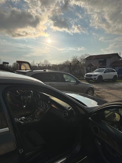 The image depicts a partially obscured view of several vehicles, predominantly Mercedes-Benz models, parked near what appears to be a rural or semi-rural setting.

Foreground: The interior of a black Mercedes-Benz vehicle dominates the immediate foreground. The open driver-side door reveals the dashboard, steering wheel, and leather seats.
Midground: Two vehicles are visible behind the foreground car. One is a silver sedan, likely a Mercedes-Benz C-Class or similar, with its front passenger door open. Further back, a white Mercedes-Benz SUV, possibly a GLC model, is parked.
Background: Behind the cars, a dilapidated building with a tiled roof is visible, suggesting an older, possibly abandoned, structure. Bare trees and utility poles further define the landscape.
Sky: The sky is partly cloudy with the sun shining through, creating a bright and slightly hazy atmosphere.

The scene seems to be situated in a village or rural area, possibly in Bohot, Bulgaria, as indicated in the prompt. The location is likely a residential or parking area with a mix of modern vehicles and older structures. There are no people immediately visible in the scene.