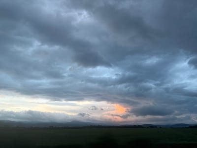 The image displays a sky filled with predominantly gray clouds, with some areas showing lighter, white-gray tones. In the upper-middle area, there's a patch of orange-yellow light breaking through the clouds. A dark strip, possibly representing land, is visible at the very top. Based on the location (Lesnovo, Bulgaria), this dark strip could be a horizon line or a land formation, obscured by distance or lighting conditions. There are no people, subjects, objects, actions, or interactions depicted in the provided image, beyond the natural elements of clouds and potentially land. The overall environment is atmospheric and suggestive of a cloudy day or impending weather.
