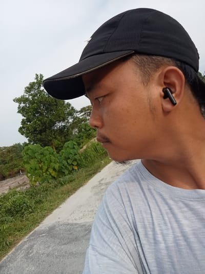 A medium shot captures a man from the neck up, facing left in profile. He wears a black baseball cap and a light blue t-shirt. A black wireless earbud is visible in his right ear. His skin tone is tan, and he has a mustache and a goatee. The background features lush green vegetation and trees, with a dirt path or road running diagonally from the lower left to the upper right. The sky is overcast. The scene appears to be outdoors in a rural or suburban area, potentially Kualu, Indonesia, given the foliage and terrain.