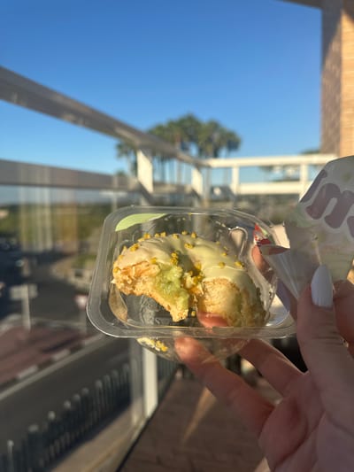 A person is holding a clear plastic container with a partially eaten pastry inside. The pastry is covered in a light green glaze and sprinkled with yellow-orange granules. The person's hand, with white nail polish, is visible. The background shows a building with large windows reflecting the blue sky and trees. The location appears to be outdoors in an urban setting. The scene is not in Beniflà, Spain.
