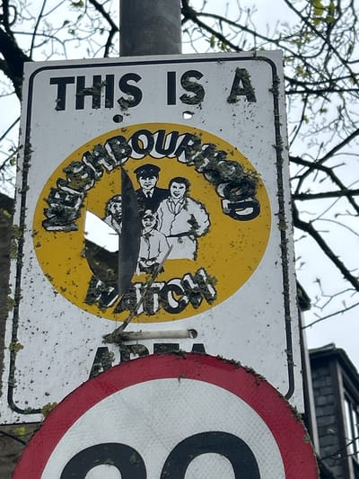 The image shows two adjacent road signs in Aberdeen City, United Kingdom. The sign on the left states, "THIS IS A NEIGHBOURHOOD WATCH" around a yellow circle containing a cartoon-style image of a group of people. The sign is visibly aged and has a hole in the center. Lichen and other biological growth are present on the signs. The sign on the right, partly visible, is a circular road sign with a red border, partially obscuring its content. Bare tree branches are visible in the background, suggesting a possible outdoor setting. The sky appears overcast. The entire scene appears to be situated outdoors in a natural environment.