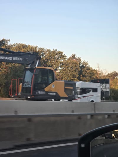 An excavator with the branding "REINHARD WEISS" and "VOLVO" is transported on a trailer along a highway. The excavator is yellow and black with orange safety railings. Adjacent to the excavator is a white recreational vehicle with a black and red stripe design. Both vehicles are positioned on a trailer and are moving along the road. The scene appears to be set during the daytime with a clear sky and trees lining the side of the highway. The highway barrier is visible in the foreground, and the reflection of a car's side mirror shows another vehicle on the road. Based on the available information, this scene is located in Bischofsheim, Germany.