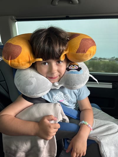 A young child with brown hair and bangs is sitting in a car seat, facing the camera. They are wearing a light blue t-shirt with a floral design and grey sweatpants. The child is leaning against a grey neck pillow with a penguin embroidered on it and a yellow travel pillow with brown spots, resembling a giraffe. Their right arm is resting on a light grey blanket, and they are wearing a colorful beaded bracelet on their left wrist. The child is smiling and looking directly at the camera. The background shows a car window with a sunshade, and a blurred view of the outdoors, suggesting the car is in motion. The scene appears to be a candid shot of the child during a car journey. This image was taken in Enaberdino, Russia.