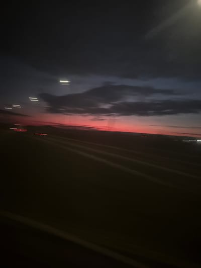 The image depicts a dark, blurry scene taken at night or twilight, possibly from inside a vehicle. A streak of red/orange light bisects the image horizontally, suggesting a sunset or city lights reflecting off clouds. The left side shows dark cloudy skies with some lighter areas. On the right, the view is obscured by dark panels, perhaps part of a car interior. There are some indistinct bright spots that appear to be light sources or reflections. The location, Belyi Rast, Russia, is not explicitly evident from the image content but may be suggested by the context in which the image was captured. No people, interactions, or specific objects are discernible.