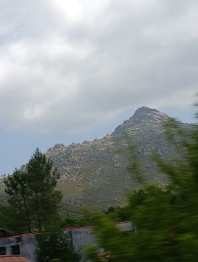 The image depicts a mountainous landscape with a prominent peak in the background, under a cloudy sky. The mountain is covered in rocks and vegetation, with trees in the foreground. A building structure with a red roof is partially visible at the bottom left of the frame. The image appears to be taken from a moving vehicle due to the motion blur on the vegetation. This scene is consistent with the natural beauty of the Peneda-Gerês National Park, located in the municipality of Terras de Bouro, Portugal.