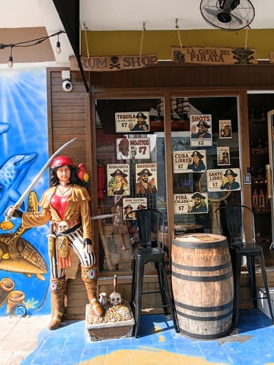 This image depicts the exterior of a rum shop in Puerto Plata, Dominican Republic. The shop appears to be themed around pirates, with a prominent wooden sign reading "LA CUEVA DEL PIRATA" (The Pirate's Cave) and "RUM SHOP" featuring a skull and crossbones. Various advertisements for drinks like Tequila, Mojitos, Daiquiri, Cuba Libre, Pina Colada, and Santo Libre are displayed on the glass storefront, with prices ranging from $3 to $20.
A life-sized pirate figurine, dressed in a brown and red outfit with a sword, stands outside the shop, adding to the pirate theme. Behind the figurine and the shop entrance, a large wooden barrel sits on the ground, possibly used for decoration or as a display. The ground in front of the shop is painted with a vibrant blue mural featuring a dolphin and a sea turtle, suggesting a coastal or tropical location. A ceiling fan is visible on the left side of the image, and a security camera is mounted on the wooden wall. The overall impression is a lively and thematic establishment catering to tourists interested in rum and a pirate-inspired experience. The time of day appears to be daytime, with bright sunlight casting shadows.
