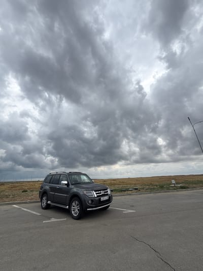 A dark gray Mitsubishi Pajero SUV is parked in a parking lot. The vehicle is positioned at an angle, facing slightly to the left of the frame. The sky above is filled with dramatic, heavy gray clouds, suggesting an overcast or stormy day. In the background, there is a flat, dry, grassy field stretching to the horizon. A line of tall poles with lights are visible in the distance on the left side of the image. A street light is present on the right side of the frame. The parking lot surface is asphalt and shows some wear and tear, including a visible crack. The location is identified as Batal'nenskoe rural settlement, Ukraine. There are no people visible in the image.