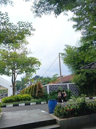 The image captures an outdoor scene, likely a pathway or open area within a campus or a well-maintained public space, filled with lush greenery under an overcast sky.
In the immediate foreground, dark grey paving stones are visible, possibly forming steps or a platform leading down. To the right of this, a large, dark blue plastic barrel stands next to a small garden area. This garden is edged by a low black fence with arched top sections, and behind it are green plants with hints of red flowers.
The midground features a paved pathway that runs horizontally across the frame. Its edge is marked by a curb painted with alternating black and white stripes. Two young women are walking from left to right on this path. Both are wearing what appears to be uniforms, consisting of light brown long-sleeved tops and dark bottoms. They both have dark head coverings (hijabs). The woman in front carries a light-colored bag, while the one behind her carries a darker bag.
Behind the pathway, a row of neatly trimmed evergreen bushes and small trees with reddish-brown foliage creates a vibrant line of color. Further back, a low wall made of black blocks with white mortar lines forms a boundary. A tall, light-colored utility pole with wires extends upwards from behind this wall on the right side of the image.
To the far left of the image, a large tree with abundant green leaves stands prominently. Partially visible behind it is a white building or structure with intricate, ornate gate details, suggesting an entrance. The upper part of the image is dominated by the bright, cloudy sky, with the canopies of several trees, their leaves illuminated by the ambient light, framing the top and sides of the view. In the distance, above the wall and behind the utility pole, the terracotta rooftops of residential buildings are discernible amongst the trees. The overall impression is a peaceful, well-maintained environment during daylight hours. No discernible text is visible in the image.