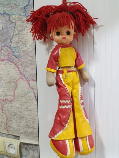 A full-body, slightly angled, close-up shot shows a vintage doll hanging on a wall next to a map of Armenia. The doll has bright red yarn hair, big black eyes, and a small pink mouth. She is wearing a red and yellow outfit with a yellow top and red pants that flare out at the bottom, with white trim. The doll's left arm is visible and appears to be made of fabric. To the left of the doll is a section of a map showing geographical and political boundaries, with names of places visible in a small font. Below the map, a white electrical outlet with two sockets is mounted on the wall. The background to the right of the doll is a vertical white surface with subtle lines, possibly a door or a wall panel. The lighting in the scene is soft, casting a faint shadow of the doll on the white surface. There are no people visible in the image, and the doll is the sole subject of focus. The scene appears to be indoors, likely in a residential or educational setting given the presence of a map and electrical outlets. The doll is positioned centrally in the frame, with the map occupying the left side and the white surface the right.