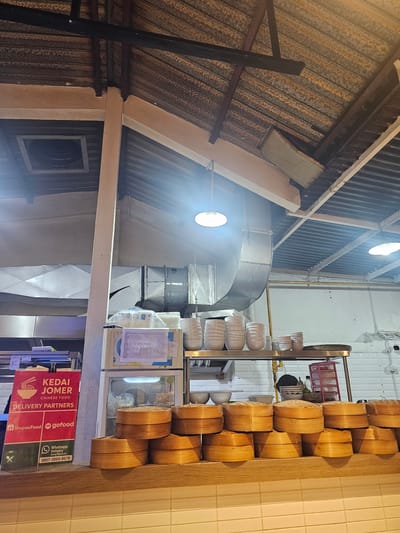 The image shows the interior of a restaurant kitchen in Parakan, Indonesia. A stack of bamboo steamers are prominently displayed on a wooden counter in the foreground. Behind the steamers, a refrigerator is visible with a sign attached to it. A red sign with "KEDAI JOMER CHINESE FOOD" and delivery partner logos such as ShopeeFood and GoFood is positioned to the left of the refrigerator. Further back, metal shelving units hold stacks of white ceramic bowls and plates. Industrial ventilation ducts are visible running along the ceiling, which is constructed of corrugated metal. A bright overhead light illuminates the space. The overall environment appears to be a functional, operational kitchen.