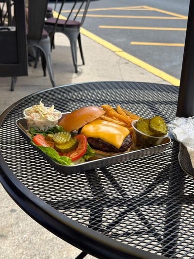 A close-up, eye-level shot shows a burger, french fries, coleslaw, and pickles served on a metal tray on a black metal picnic table. The burger features a golden bun, a beef patty, and a slice of melted yellow cheese. Lettuce, tomato slices, and pickle slices are arranged beneath the burger. The french fries are stacked behind the burger, and the coleslaw is in a small clear plastic cup. A small cup of pickle slices is also on the tray. In the background, yellow lines are painted on asphalt, suggesting a parking lot or street. Chairs with dark cushions are visible behind the table, and a dark pillar stands to the right. The lighting suggests an outdoor setting on a sunny day, casting shadows on the table. The location is identified as Edgewater, United States of America.