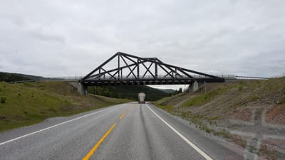 This image shows a highway passing under a large, dark-colored metal truss bridge. The bridge spans the highway at an angle, with its supports built into the sloped, grassy embankments on either side of the road. The road itself is asphalt with white and yellow lane markings. A white truck with a large trailer is driving away from the viewer, under the bridge. The surrounding landscape is hilly and covered with green grass and trees, with a line of darker green trees visible in the distance. The sky is overcast and cloudy, suggesting a cool or temperate climate. The overall scene is one of a rural highway infrastructure in a natural setting. The location is identified as Trofors, Norway. There is no visible text in the image.