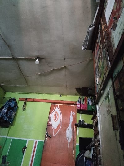 The image shows the interior of a room located at coordinates -7.545 latitude and 112.276 longitude in Kecamatan Peterongan, Indonesia. The room is sparsely furnished and features a light brown wooden door with a white outline of a bird and a large white abstract shape painted on it. The wall is painted green, and the ceiling is gray. A white light bulb is visible in the center of the image. There is a shelf on the right wall, holding a variety of objects, including several folders. A blue cloth garment is hanging on a hook on the left wall. The environment appears to be indoors, possibly a residential home.