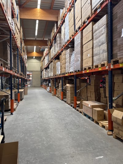 The image shows the interior of a large warehouse with high ceilings and concrete floors. Tall metal shelving units are filled with numerous cardboard boxes of various sizes, some wrapped in plastic. The shelves are organized in parallel aisles, creating a perspective view down the length of the warehouse. Lighting fixtures are visible on the ceiling, illuminating the space. Wooden beams and a metal roof structure are also apparent. In the foreground, a hand pallet truck with cardboard boxes on its forks is positioned on the left side. The overall environment suggests a storage or distribution facility. The image does not contain any people or specific identifiable landmarks that would allow for precise location within Bad Säckingen, Germany.