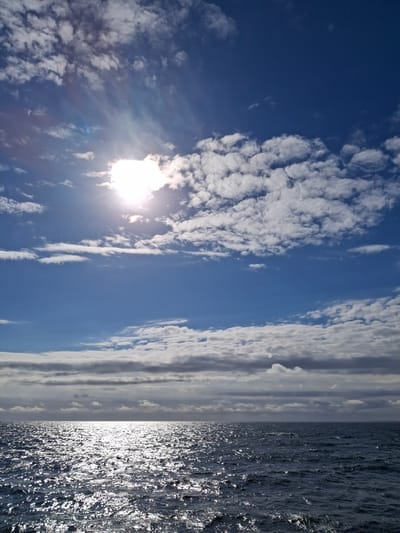 The image shows a view of the sky and ocean, likely taken from a boat or the shore. The left side of the frame is filled with a blue sky dotted with cumulus clouds. The sun is visible, causing a bright glare. The right side of the image features the ocean, its surface rippled and reflecting sunlight. The location is given as the city of Unknown, Denmark. No people or other subjects are visible.