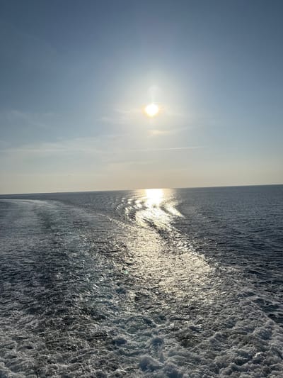 The image depicts a maritime scene, observed from the stern of a moving vessel, showing its wake on the open sea near Kamariotissa, Greece. The bright, undiffused sun is positioned in the upper center-right quadrant of a clear, light blue sky, casting an intense, shimmering reflection across the water surface, extending from the horizon towards the foreground. A distinct, foamy white wake trails from the bottom right corner, curving towards the left mid-ground, indicating the vessel's propulsion through the dark blue-gray water. Faint cirrus clouds are visible, and a single, thin contrail traverses the sky horizontally in the upper mid-section. No individuals, other vessels, or landmasses are visible in the frame.