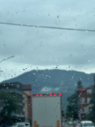 The image, taken through a rain-streaked window, depicts a view of Dubove, Ukraine on a cloudy day. In the foreground, numerous raindrops distort the scene. A blurred white truck with four red lights on its rear is visible in the mid-ground, facing away from the camera. To the left of the truck, a multi-story building with dark windows can be seen. To the right, a building with red roofing and white windows is partially obscured by trees. In the background, a mountain range is silhouetted against the overcast sky. The overall impression is one of a damp, overcast day, likely in the late afternoon or early evening given the red lights on the truck.