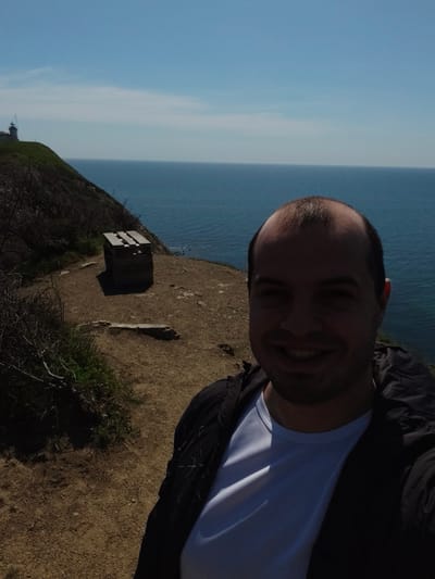 A medium shot of a man smiling while taking a selfie. He is wearing a white t-shirt and a black jacket. Behind him is a dirt path with a wooden structure on the left. In the distance, there is a lighthouse on a cliff, and beyond that, the sea and sky are visible. The location is Emona, Bulgaria.