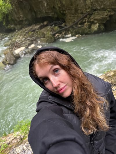 A person with light skin and long, light brown, wavy hair, wearing a black hooded jacket, takes a selfie in Tsabal, Abkhazia. The background shows a river or creek flowing through a rocky, moss-covered canyon.
