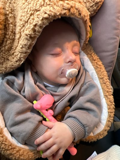 A sleeping infant is centrally positioned in a medium close-up, appearing to be in a car seat or stroller in Oberstenfeld, Germany. The infant's eyes are closed, and a clear pacifier with a white, rainbow-patterned disc is present in its mouth. A minor reddish mark is visible on the infant's forehead. The infant is dressed in a grey long-sleeved hooded top and covered by a textured, light brown, plush hooded garment with a white lining. The infant's hands are clasped around a pink and lime green teething toy, shaped abstractly. The background consists of a grey upholstered surface, consistent with a child safety seat or vehicle interior.
