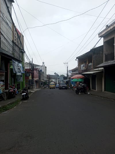 This image depicts an overcast street scene in Houaythat, Laos, captured from a low vantage point looking down a paved, two-lane road that curves slightly to the right in the distance. The sky is uniformly light grey.
On the left side of the street, multi-story buildings with varied facades line the sidewalk. The nearest building is predominantly grey concrete with numerous black electrical cables running vertically along its exterior. A green awning and a blue banner are affixed to its lower section. Two motorcycles, one red and one black, are parked on the sidewalk near this building, which also shows signs of weeds growing from cracks. Further down the street on the left, other buildings display colors like pink, white, and various shades of grey, with exposed brick or concrete textures. A white building in the mid-distance features a small, minaret-like tower on its roof.
The right side of the street is also bordered by multi-story buildings. The closest building has a light brown upper facade with a balcony enclosed by white railings, and a lower section with dark, stone-like textured cladding. A shop with a dark green roll-down shutter is visible beneath it. Further along this side, two large, colorful umbrellas, one orange and one green, are set up on the sidewalk, partially obscuring a person standing beneath the green umbrella, facing away from the camera. A dark motorcycle is parked nearby. Another dark-colored SUV is parked on the road further down on the right.
The asphalt road surface appears dry and displays some minor texture variations. Overhead, a dense network of black electrical and communication wires crisscrosses the scene, connecting to power poles and buildings. In the mid-distance, a yellow van is visible on the left side of the road, followed by a dark car and a light blue car. Another individual is partially visible riding a scooter further down the street on the left. The general environment suggests a bustling commercial or residential area.
