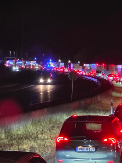 The image depicts a nighttime scene of a traffic jam on a highway, likely in or near Uettingen, Germany, evidenced by the German license plate "MN IH 194" on the car in the foreground. Several lanes of vehicles, including trucks and cars, are stationary. Emergency lights, flashing blue and red, are visible on some vehicles, suggesting a potential accident or road closure ahead. The road surface appears wet, reflecting the vehicle lights. The surrounding environment includes grassy verges and roadside vegetation. A circular sign with a white symbol on a dark background is present on a pole in the mid-ground. The overall atmosphere is dark, illuminated primarily by the artificial lights of the vehicles and emergency services.