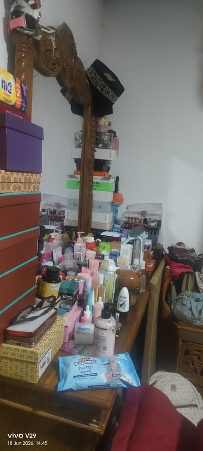 The image depicts a cluttered dresser surface and surrounding area, likely in a bedroom, located in the city of Pulodolang, Indonesia. The dresser is covered with an assortment of personal care products, including various bottles, jars, and tubes of lotions, creams, and other toiletries. Some items are stacked precariously on top of each other, creating a disorganized appearance.
Prominent objects include several stacked boxes, a wooden carved decorative piece, and two black kufi hats placed on top of the carved piece. Among the toiletries, there are visible brands like "Kodomo baby wipes" and "Rexona". A pair of eyeglasses rests on a patterned box to the left. In the background, there are more stacked boxes, some appearing to be electronics packaging, including "VIVO" branded boxes.
A large piece of red fabric, possibly clothing, is draped over part of the furniture on the right side of the image, partially obscuring a backpack with a repeating M K M K pattern. A blue patterned cloth bag is also visible on a wooden stand to the right. The lighting suggests an indoor environment, likely with artificial light sources. The overall impression is of a lived-in space with personal items accumulated on surfaces. The timestamp "vivo V29 18 Jun 2026, 14.45" is present in the bottom left corner, indicating the time and device the photo was taken.