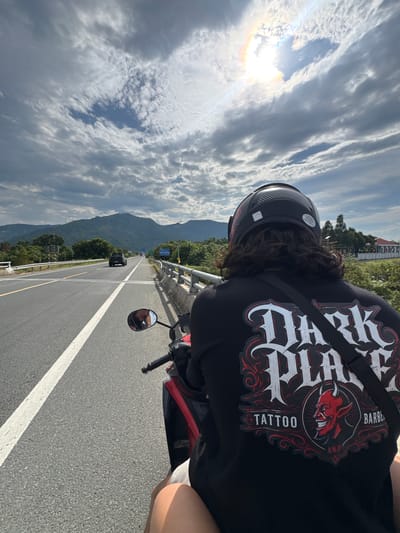A first-person perspective shot from a motorcycle on a highway shows a person wearing a black t-shirt with a red and white graphic of a devil and text that reads "DARK PLAY TATTOO AND BARBER" and a black helmet. The motorcycle is traveling on a paved road with a white line marking the lane. In the distance, there are green hills, trees, and mountains under a cloudy sky with the sun visible and casting a rainbow halo. The road appears to be in a rural area with sparse vegetation and a few buildings visible on the right side. The scene is likely in Suối Tân, Vietnam, given the geographical context provided.