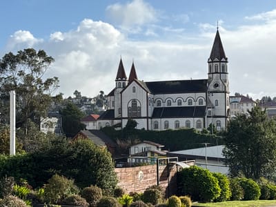 The image shows the Sacred Heart of Jesus Church in Puerto Varas, Chile, taken on a partly cloudy day. The church, a prominent white building with red-roofed spires, stands on a rise amidst lush greenery. Below, a mix of residential and commercial buildings are partially obscured by trees and vegetation. A wooden fence is visible in the foreground, bordered by manicured bushes and lawn. The sky is a mix of blue and cloudy patches.
