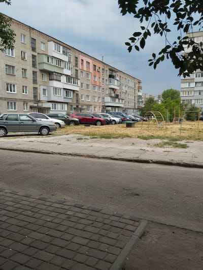 A daytime, medium shot captures a street scene in Sheptytskyi, Ukraine, featuring a row of parked cars in front of a multi-story apartment building. The building, constructed with light-colored brick, exhibits various architectural styles with differing balcony designs and colored facades on some sections. In the foreground, a paved sidewalk transitions to a street with some visible wear. Beyond the parked cars, a dry, grassy field is dotted with playground equipment, including a metal arch climber and swings. Trees are visible in the background, suggesting a partially urbanized, residential area. The sky is overcast, indicating potentially cloudy weather. No people are clearly visible, and the overall scene conveys a sense of everyday life.