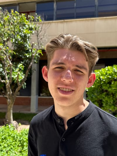 A young Caucasian male is standing outdoors. He has blonde hair, fair skin, and is wearing a black button-down shirt. He is smiling and looking at the camera. The background includes green trees and a building with blue windows. The location is Sant Vicent del Raspeig, Spain.