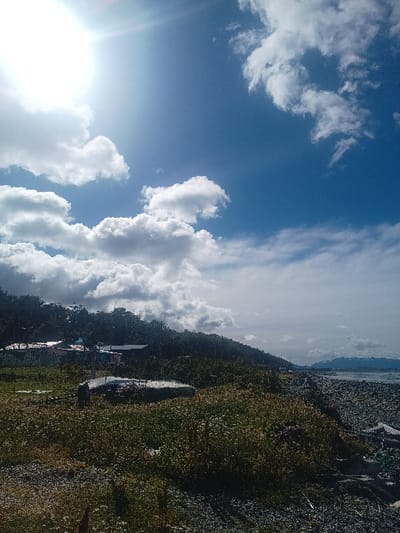 A landscape scene presents a vibrant blue sky dotted with fluffy white clouds, transitioning to a grassy terrain with hints of a gravelly path. A wooded area with buildings nestled within is visible, leading to a distant, hazy mountain range. The sun shines brightly, casting a glare across the bottom left of the frame, partially obscuring the details in that area. The image is rotated, causing the sky and ground to appear as if on their sides.