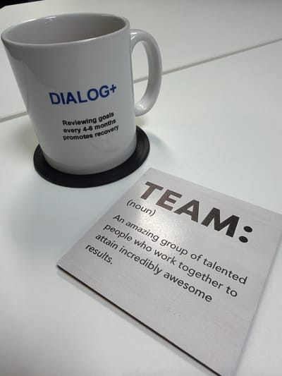 A still life scene presents a white coffee mug labeled "DIALOG+" along with the text "Reviewing goals every 4-6 months promotes recovery" on its side. The mug rests on a black circular coaster. Adjacent to the mug is a white card or coaster with the word "TEAM:" in large font, followed by the definition "(noun) An amazing group of talented people who work together to attain incredibly awesome results." The objects are positioned on a white surface. No individuals are visible in the image. There is no specific information within the image to ascertain a location in the city of Mold, United Kingdom.