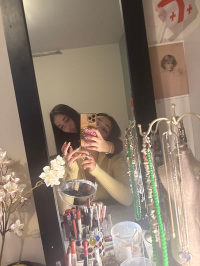 This image captures a mirror selfie featuring two young females within a private residence in Arthès, France.
**Scene Description:**
The primary visual consists of a black-framed rectangular mirror reflecting two individuals and portions of a room. On the surface in the immediate foreground, in front of the mirror, various personal items are arranged.
**Individuals:**
1. **Foreground Subject:** A female, partially visible, wearing a light-yellow long-sleeved top. Her left hand holds a gold-colored smartphone with a light-pink, textured case, positioned to capture the mirror reflection. Her fingernails are painted yellow, and she wears multiple rings on her fingers. Her eyes are directed towards the phone.
2. **Background Subject:** A female with long, dark hair is positioned behind the foreground subject. Her left arm is wrapped around the foreground subject's shoulder. Her head is resting above the foreground subject's right shoulder, with her lips pursed in a "kissing" or pouting expression. She is wearing a dark-colored garment visible on her arm.
**Objects and Environment:**
* **Mirror Reflection:** Shows a plain, light-colored wall.
* **Room Details (outside mirror's immediate reflection):**
* **Wall (right of mirror):** A small Georgian flag (white with a large red cross and four smaller red crosses in the quadrants) is affixed near the top. Below it, a framed monochrome portrait depicting a youthful individual is displayed.
* **Surface (in front of mirror):**
* To the left, a vase contains artificial light-colored flowers, primarily white and light pink blossoms.
* A small, circular cosmetic mirror is present.
* A clear, multi-compartment organizer, decorated with colorful mosaic-like pieces at its base, is filled with various makeup pencils, lipsticks, and other cosmetic tools.
* On the right, a white metal jewelry stand, resembling a small tree, holds numerous necklaces, including strands of green beads and metallic chains with pendants.
* Additional clear plastic containers are also visible.
**Lighting:** The scene is illuminated by interior lighting, casting soft shadows.