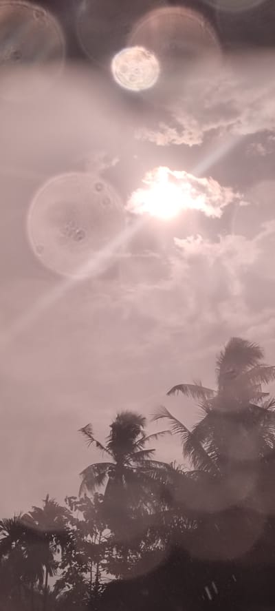 The image, taken in Matangpanyang, Indonesia, shows a sky with scattered clouds and a bright sun partially obscured by a cloud. Circular lens flares are prominent in the upper portion of the image, resembling hazy moons. The bottom portion of the image includes dark silhouettes of tropical trees, including what appear to be palm trees. The overall color tone of the image is sepia. There are no people visible.
